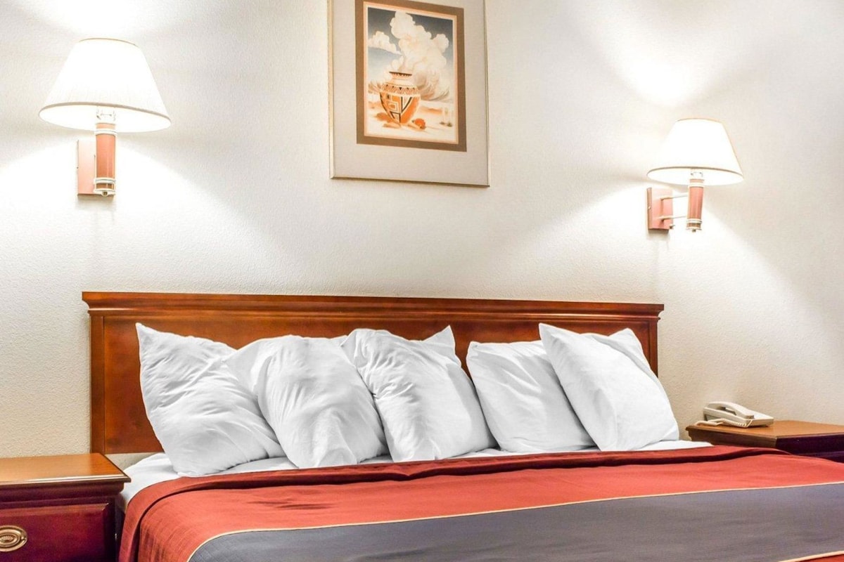 A hotel room with a wooden headboard, a bed with white pillows, a red and grey bedspread, two bedside lamps, and a framed picture on the wall.