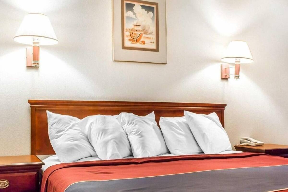 Ett hotellrum med en sänggavel i trä, en säng med vita kuddar, ett rött och grått sängöverkast, två sänglampor och en inramad tavla på väggen.