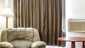 Un fauteuil beige, une table ronde en bois, un rideau à motifs, un lampadaire et un climatiseur fixé au mur.