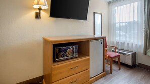 A compact room with a flat-screen TV mounted on the wall, a wooden cabinet with drawers, a small refrigerator, a chair, and a window with sheer curtains. - Rodeway Inn Asheville (Asheville)