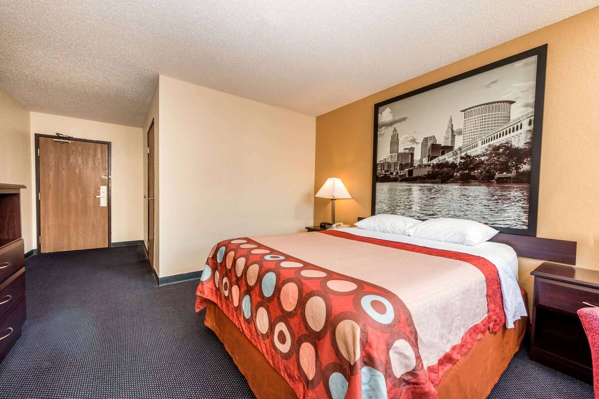 Ein Hotelzimmer mit einem großen Bett, einem Nachttisch, einer Lampe, einer Tür und einem Stadtbild an der Wand.