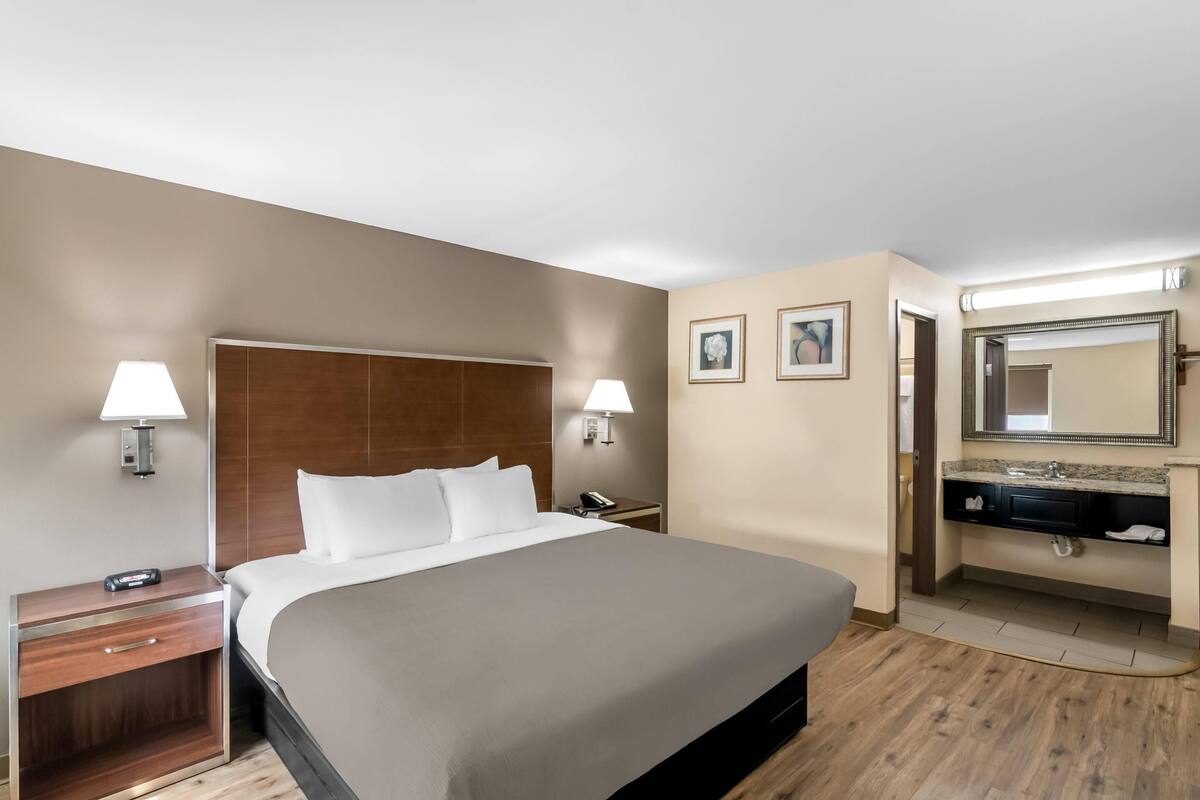 A hotel room with a large bed, bedside tables with lamps, a nightstand, a bathroom with a sink and mirror, and framed pictures on the wall.