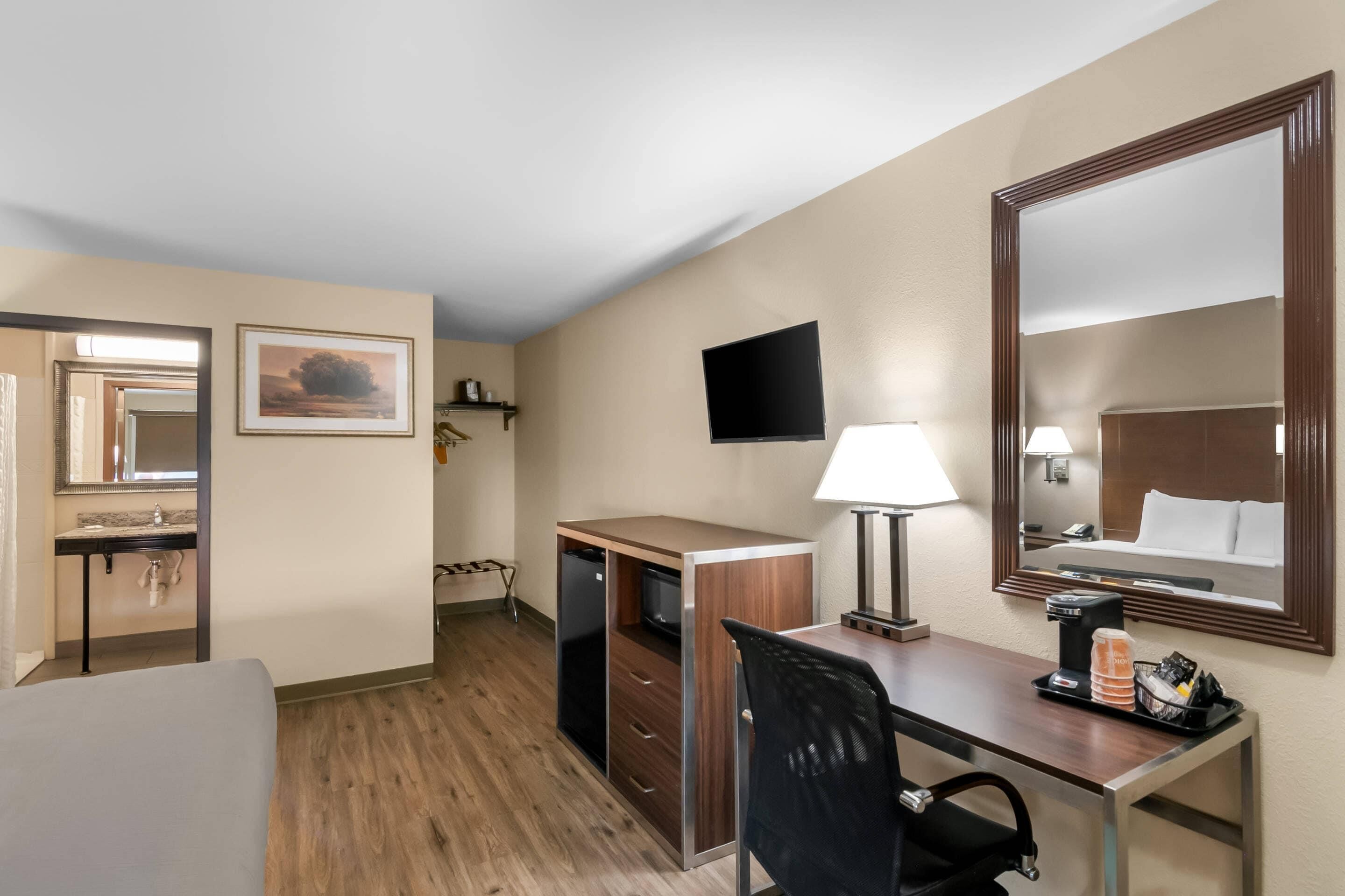 Habitación de hotel con escritorio, silla y un espejo grande. Hay un televisor montado en la pared, una lámpara y una cafetera sobre el escritorio.