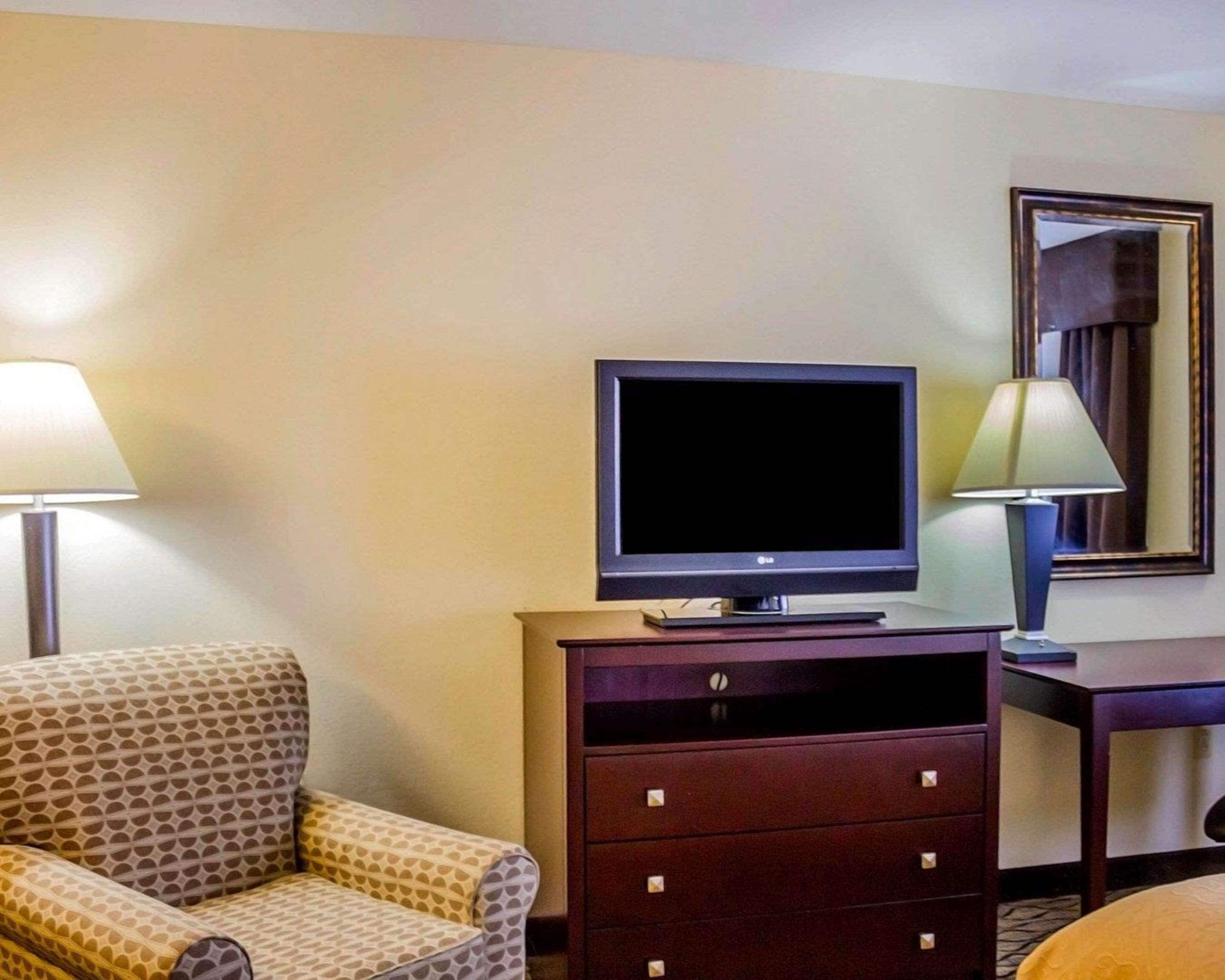 Habitación de hotel con televisión sobre un mueble de madera, un sillón estampado, un escritorio con lámpara y un espejo en la pared.