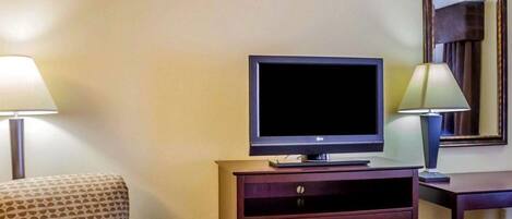 Habitación de hotel con televisión sobre un mueble de madera, un sillón estampado, un escritorio con lámpara y un espejo en la pared.