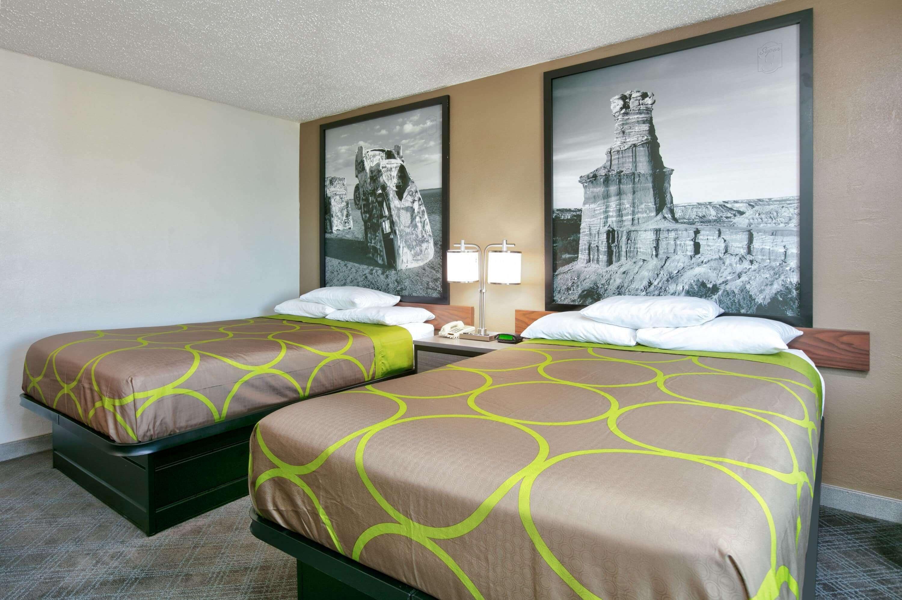 A hotel room with two single beds, a nightstand, a lamp, and two framed pictures on the wall.