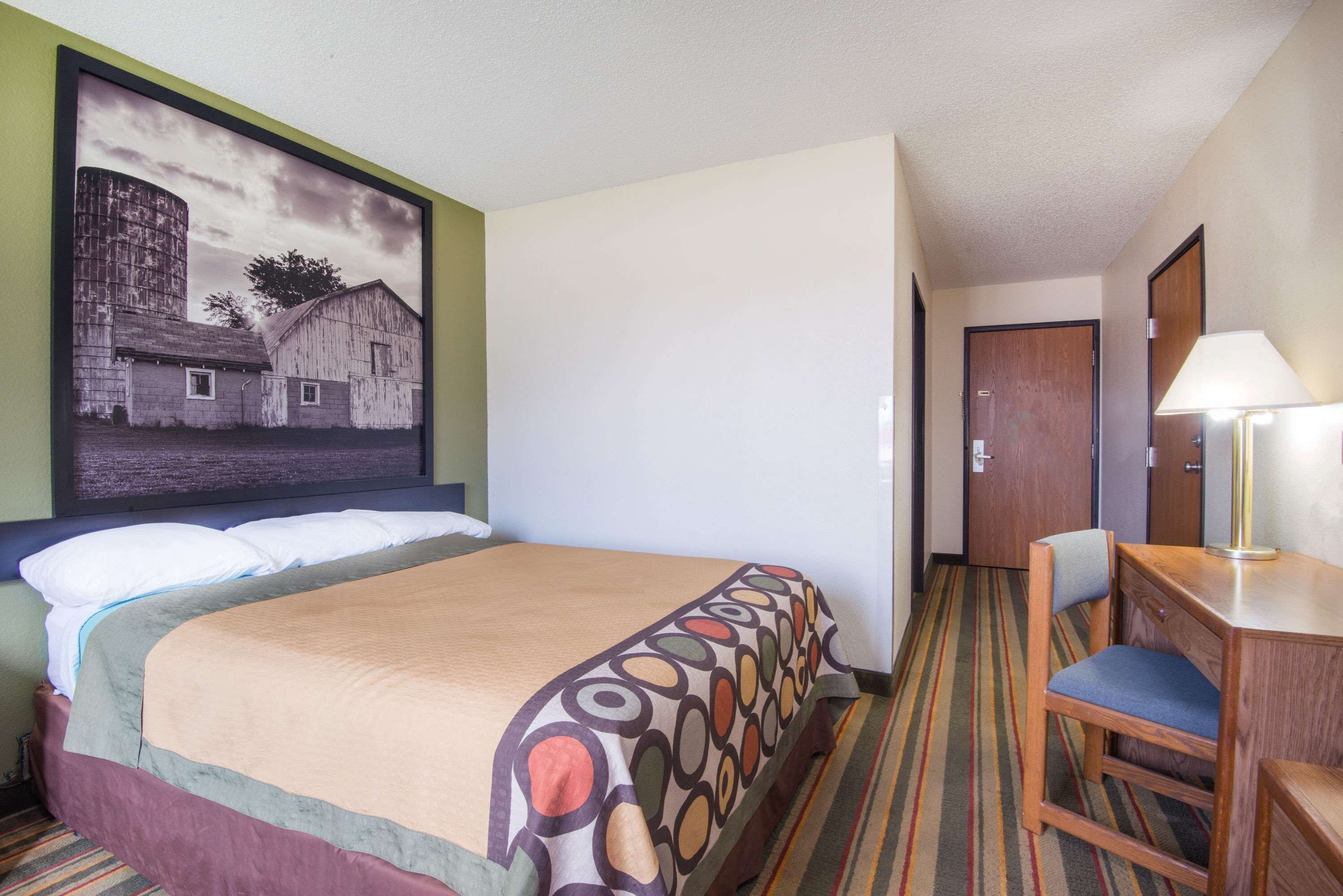 A hotel room with a bed, a desk, a chair, a lamp, and a painting on the wall.