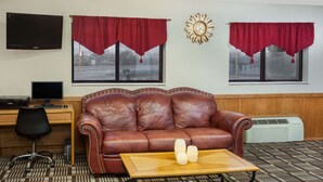 Una habitación con sofá de cuero marrón, una mesa de centro de madera con velas, un escritorio con computadora y dos ventanas con cortinas rojas.