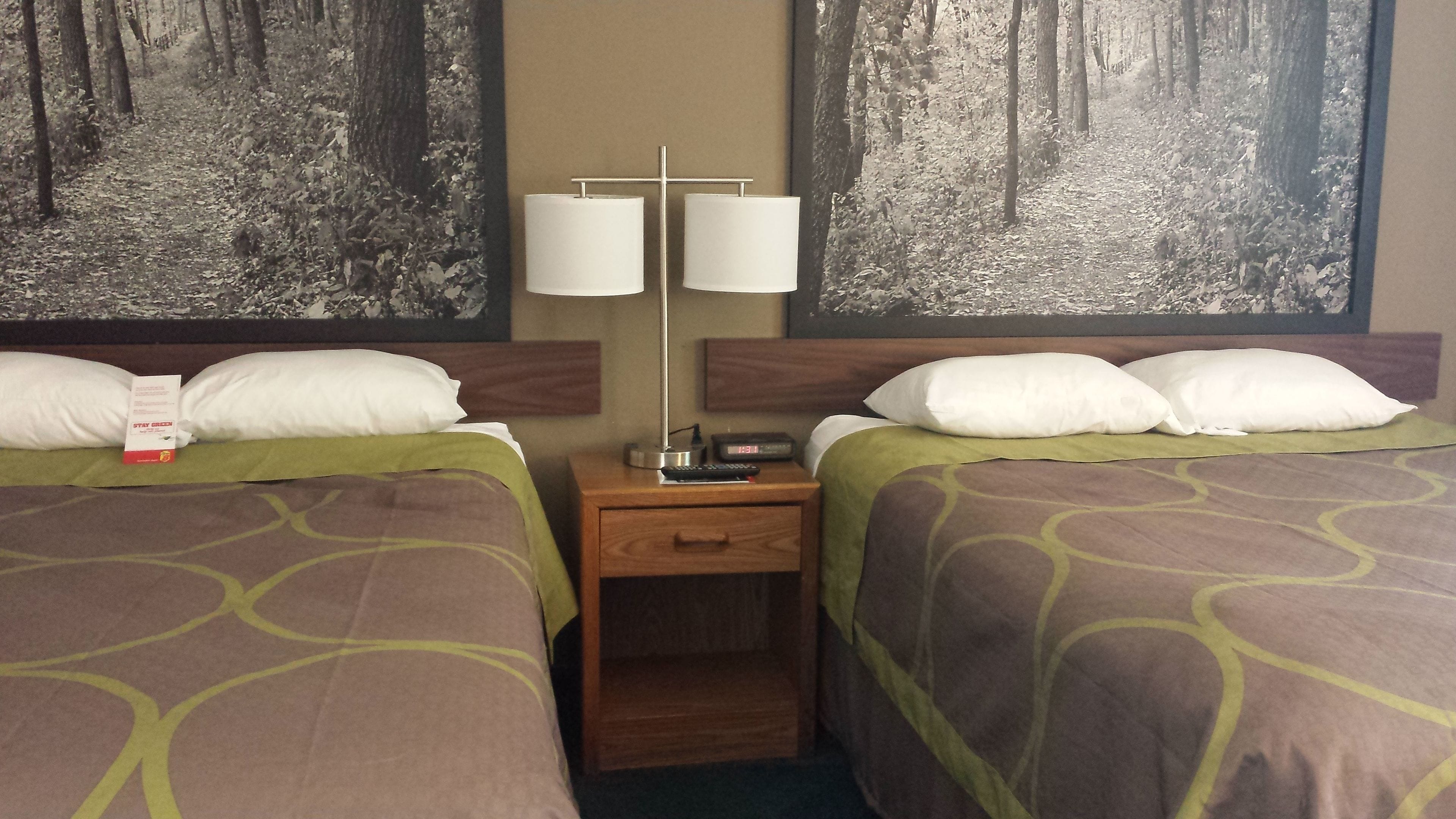 Habitación de hotel con dos camas, cada una con almohada y cubrecama estampado en tonos verdes. Se ve una mesita de noche con una lámpara, un reloj y un control remoto.