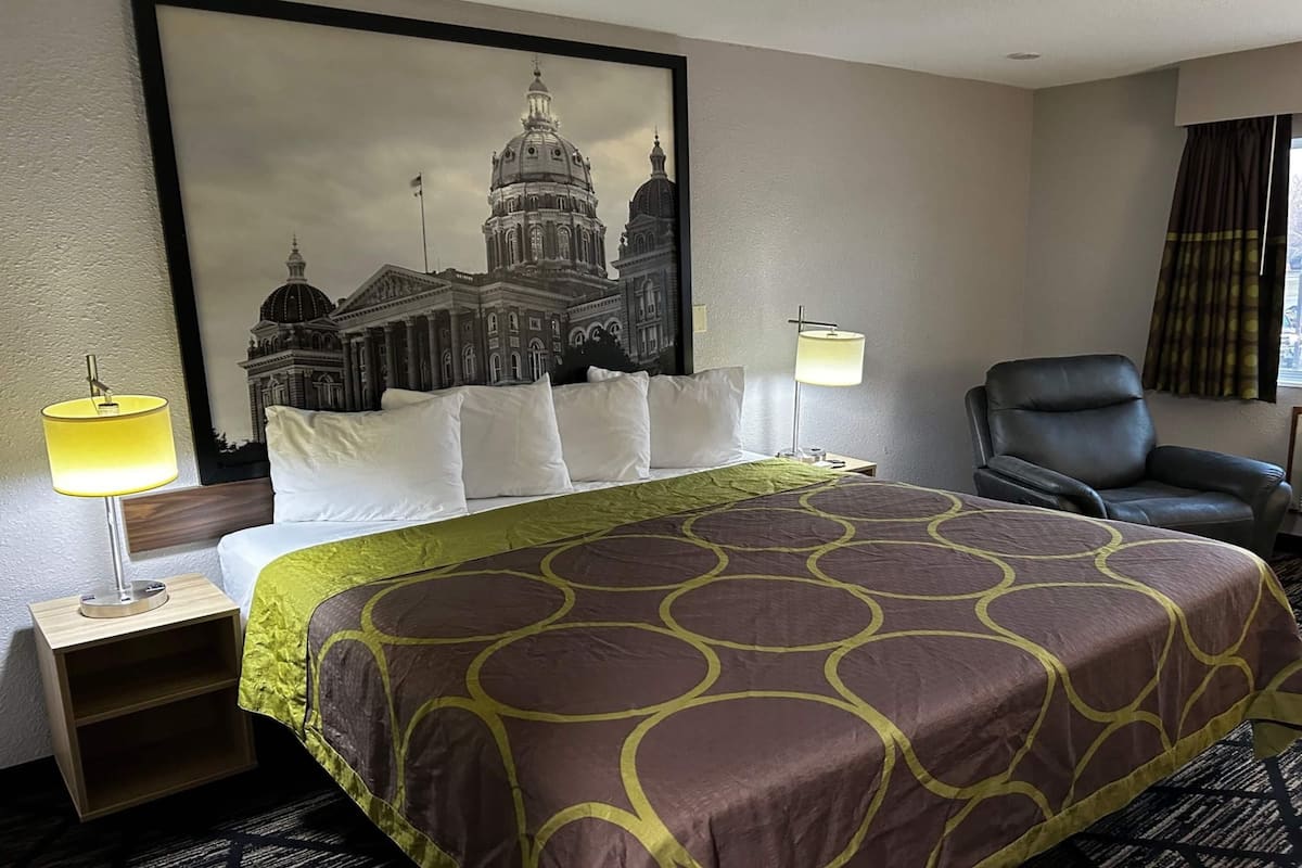 A hotel room with a large bed, two bedside lamps, a leather chair, and a framed cityscape picture on the wall.