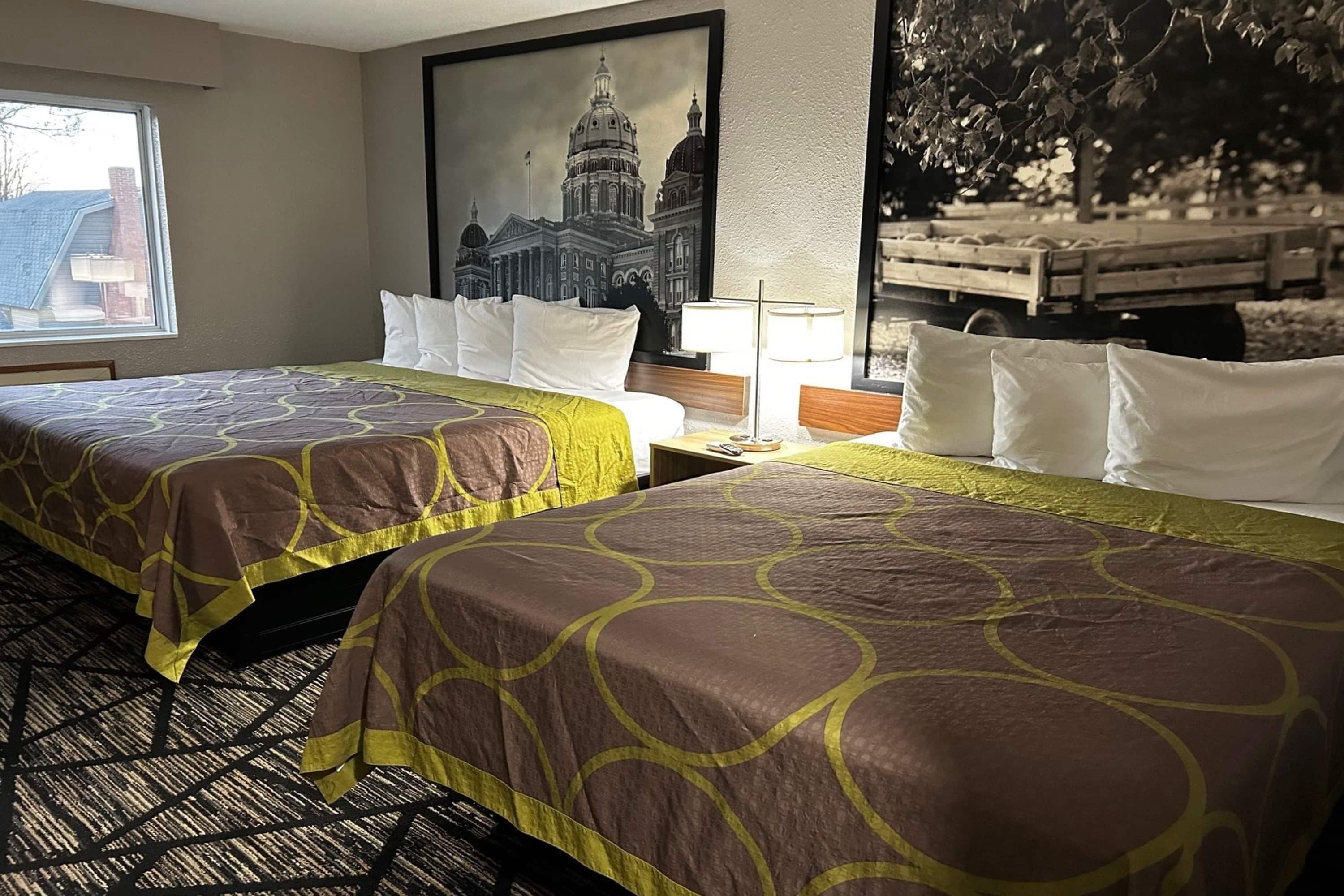 Une chambre d’hôtel avec deux lits, une grande fenêtre, un tableau encadré représentant un bâtiment et une lampe.