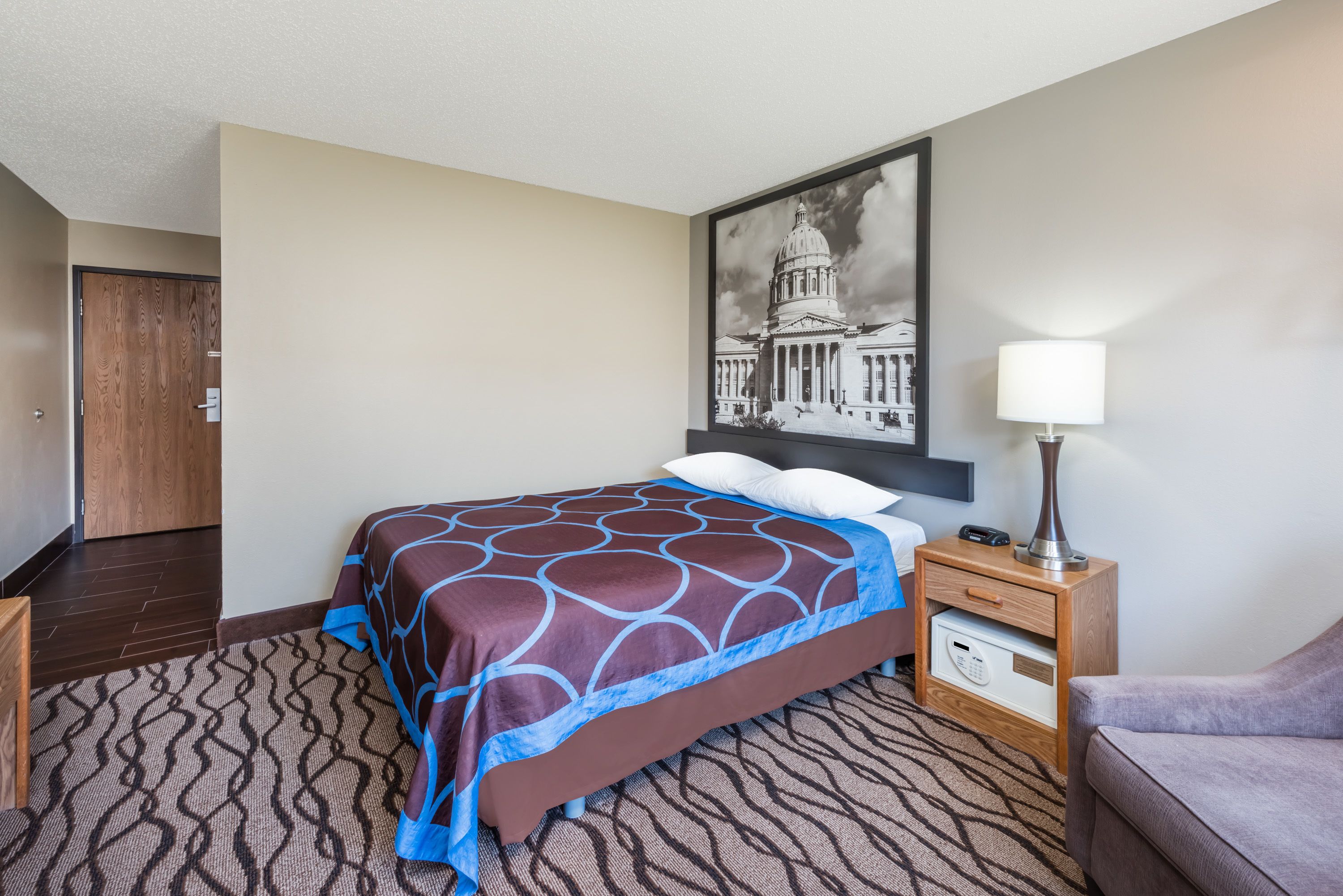 A hotel room with a bed, a nightstand, a lamp, a framed picture on the wall, and a door.