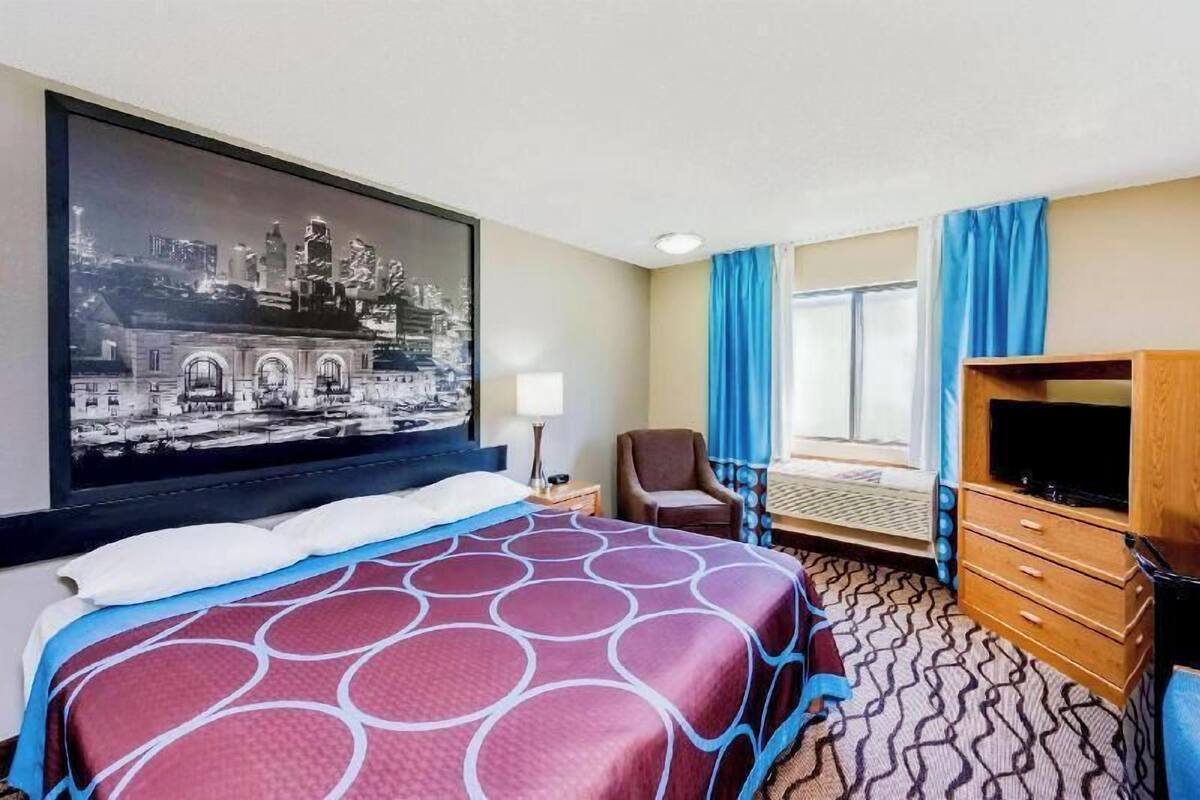 A hotel room with a bed, a television, a chair, a desk, and a large framed cityscape picture on the wall.