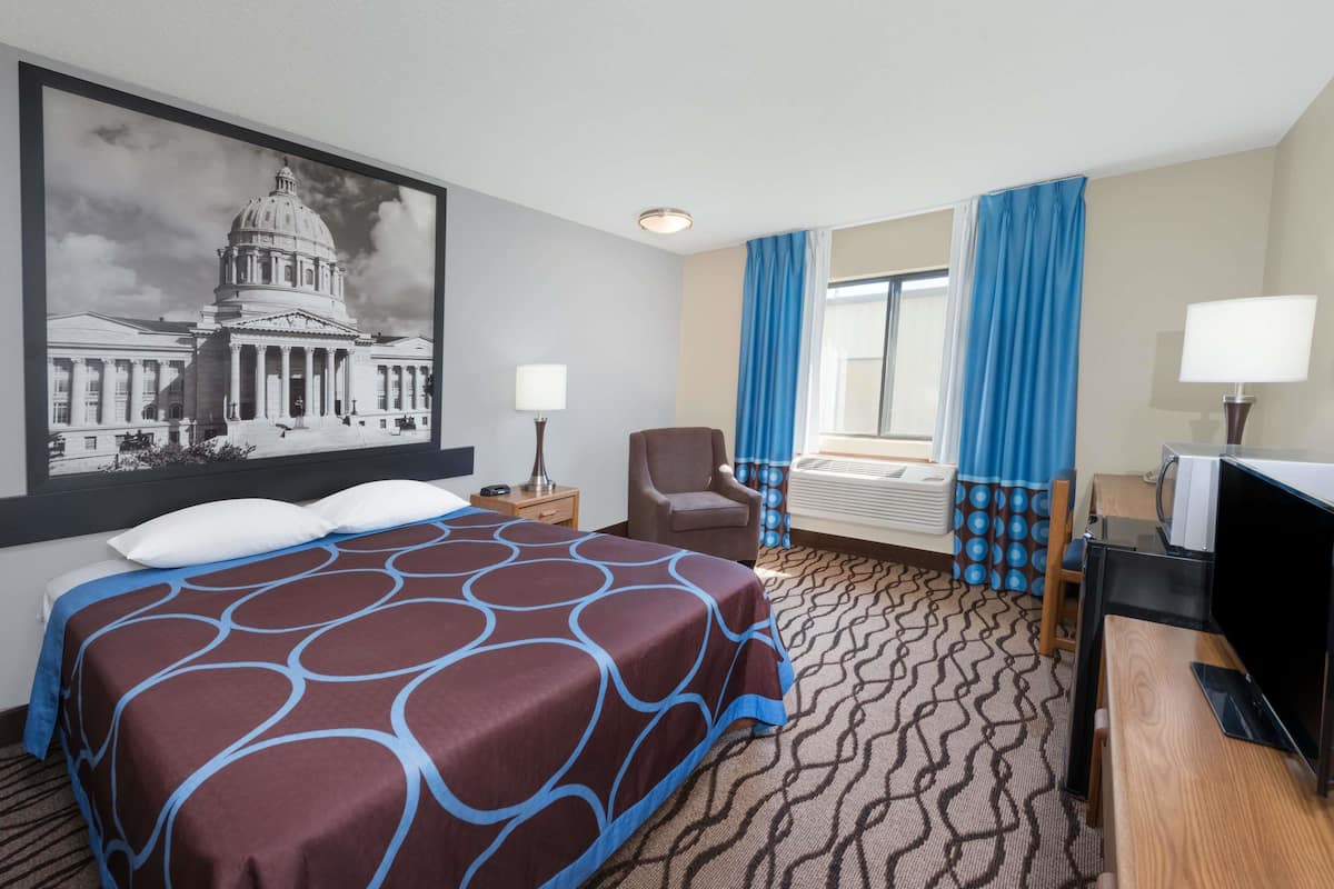 A hotel room with a large bed, a desk with a TV, a chair, a lamp, and a large window with curtains.