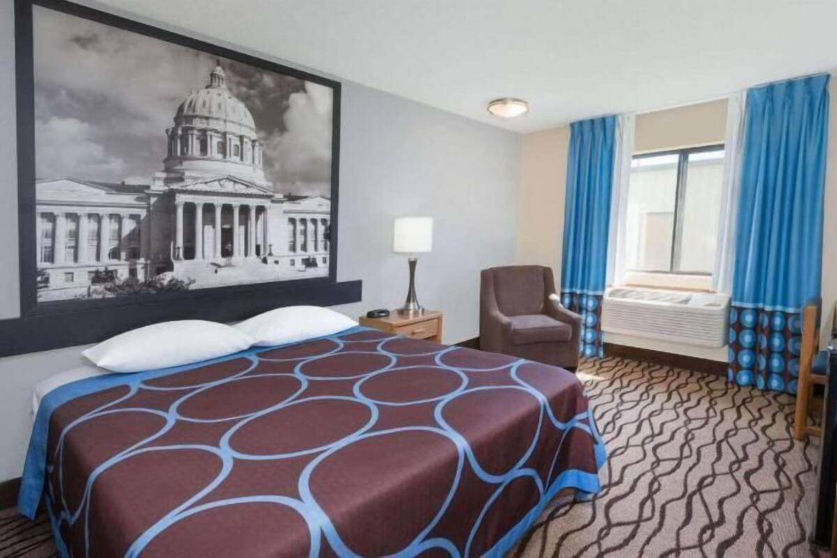 A hotel room with a large bed, a desk with a computer, a chair, a window with blue curtains, and a wall-mounted TV.