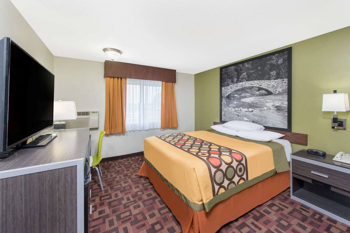 A hotel room with a bed, a desk with a TV, a lamp, a chair, and a window with curtains.