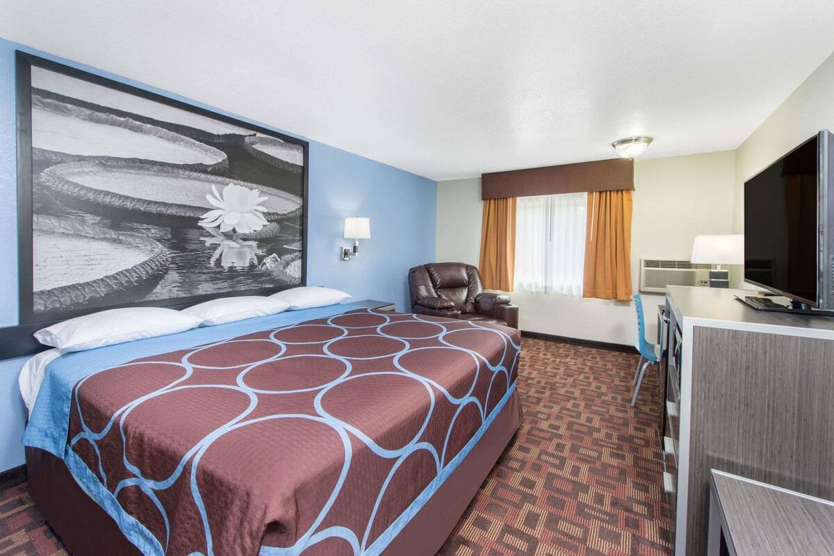 A hotel room with a large bed, a desk with a TV, a chair, and a wall-mounted lamp.