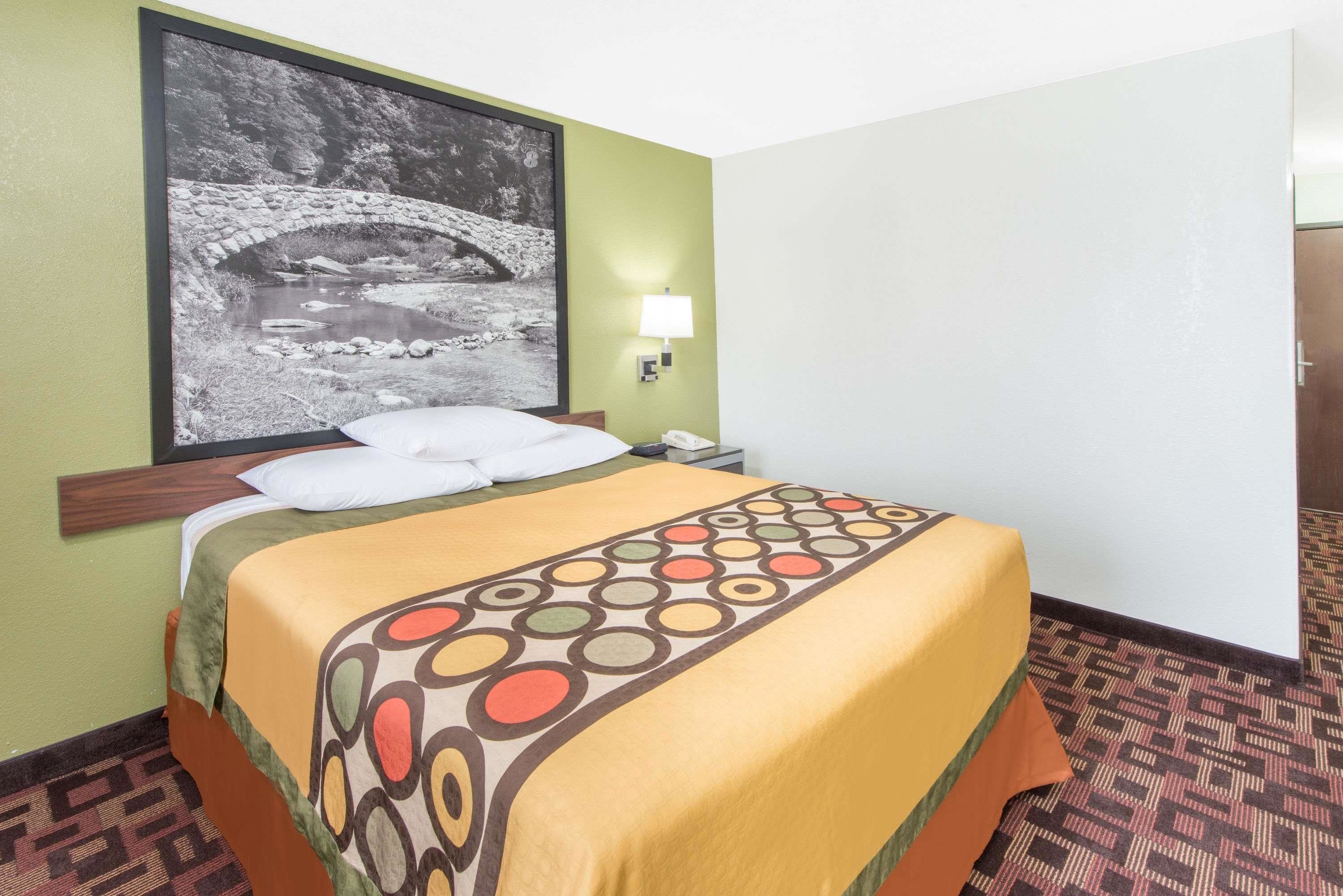 Een hotelkamer met een bed, nachtkastje, lamp en een lijst aan de muur.