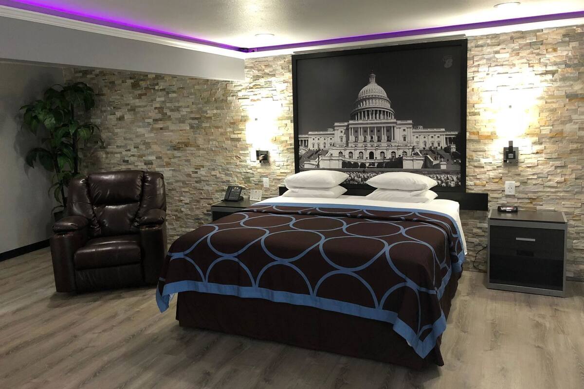A bedroom with a bed, a leather chair, a nightstand, a wall-mounted lamp, and a framed picture of a building.
