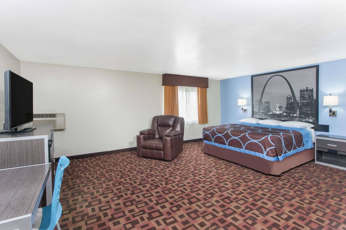 A hotel room with a bed, a desk with a television, a chair, and a cityscape painting on the wall.