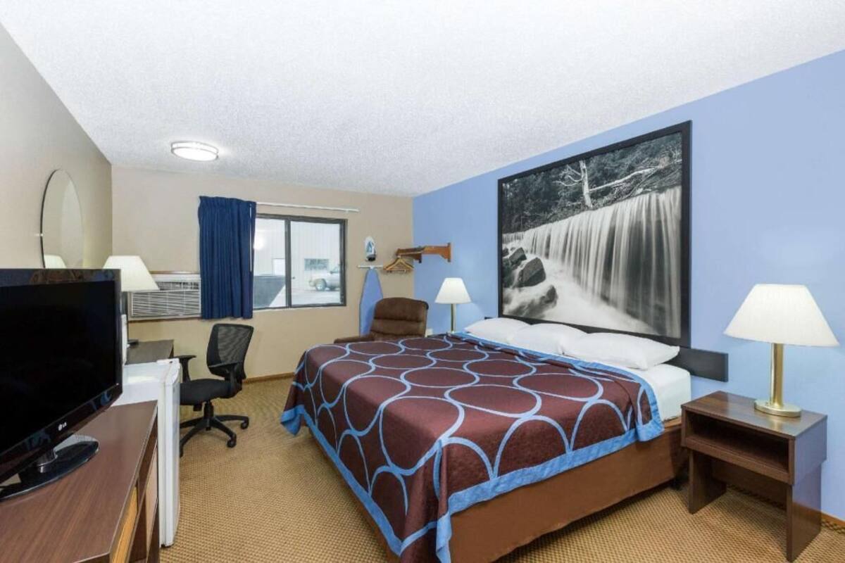 Una habitación de hotel con una cama, un escritorio con una computadora, una silla, un televisor y un ventanal grande con cortinas.