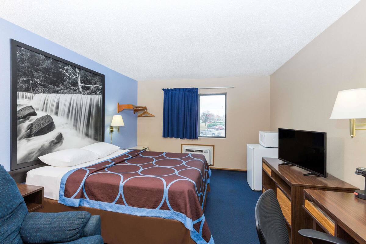 Habitación de hotel con una cama, un escritorio con televisor, una silla, una lámpara de escritorio, una ventana con cortinas azules y un calentador instalado en la pared.