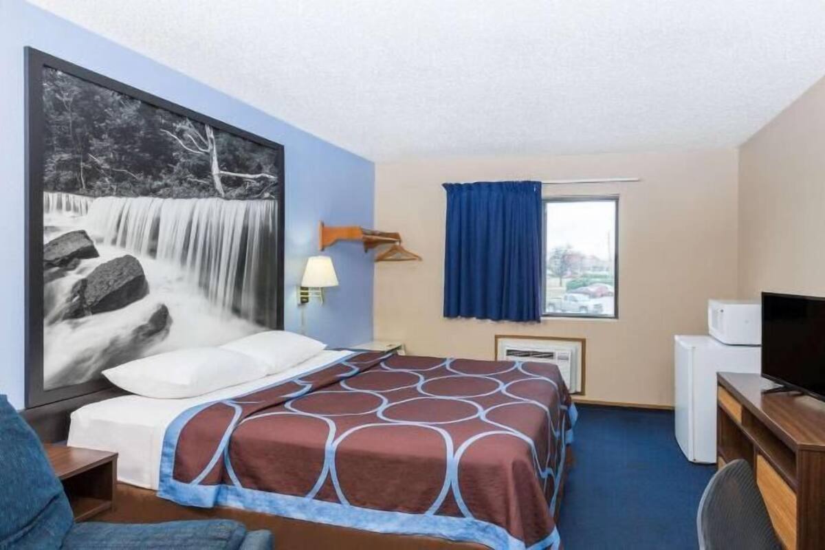 Habitación de hotel con cama, escritorio, televisor, ventana con cortinas y lámpara de pared.
