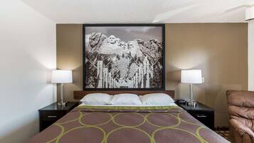 Ein Hotelzimmer mit einem Bett, zwei Nachttischlampen, einer braunen Bettdecke mit Muster, einem gerahmten Bild von einer Berglandschaft und einem braunen Ledersessel.