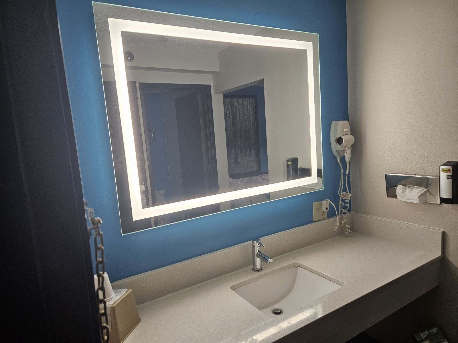 A bathroom with a large mirror, a sink, a hairdryer, and a tissue dispenser.