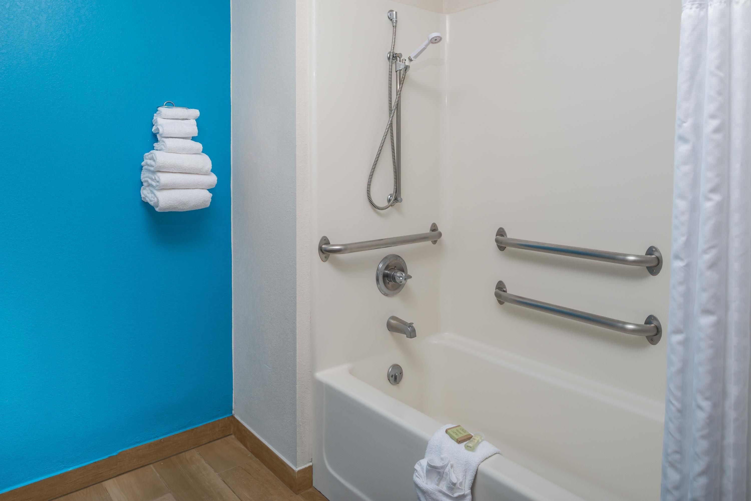 Une salle de bain avec une baignoire blanche, des barres d’appui, une pomme de douche et une pile de serviettes blanches, le tout sur un mur bleu.