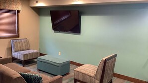 Een woonkamer met een flatscreen televisie aan de muur, een blauwe poef en twee gestoffeerde fauteuils.
