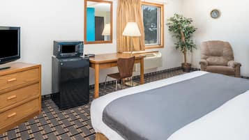 A hotel room with a bed, a desk with a computer, a TV, a lamp, a chair, a potted plant, and a clock on the wall.