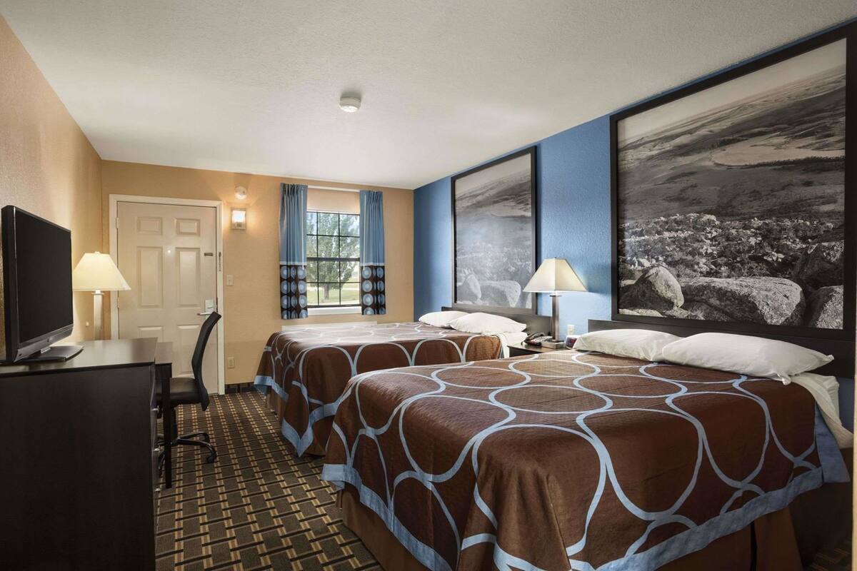 Quarto de hotel com duas camas, uma mesa de trabalho com televisão, uma cadeira, uma janela com cortinas e um quadro na parede.