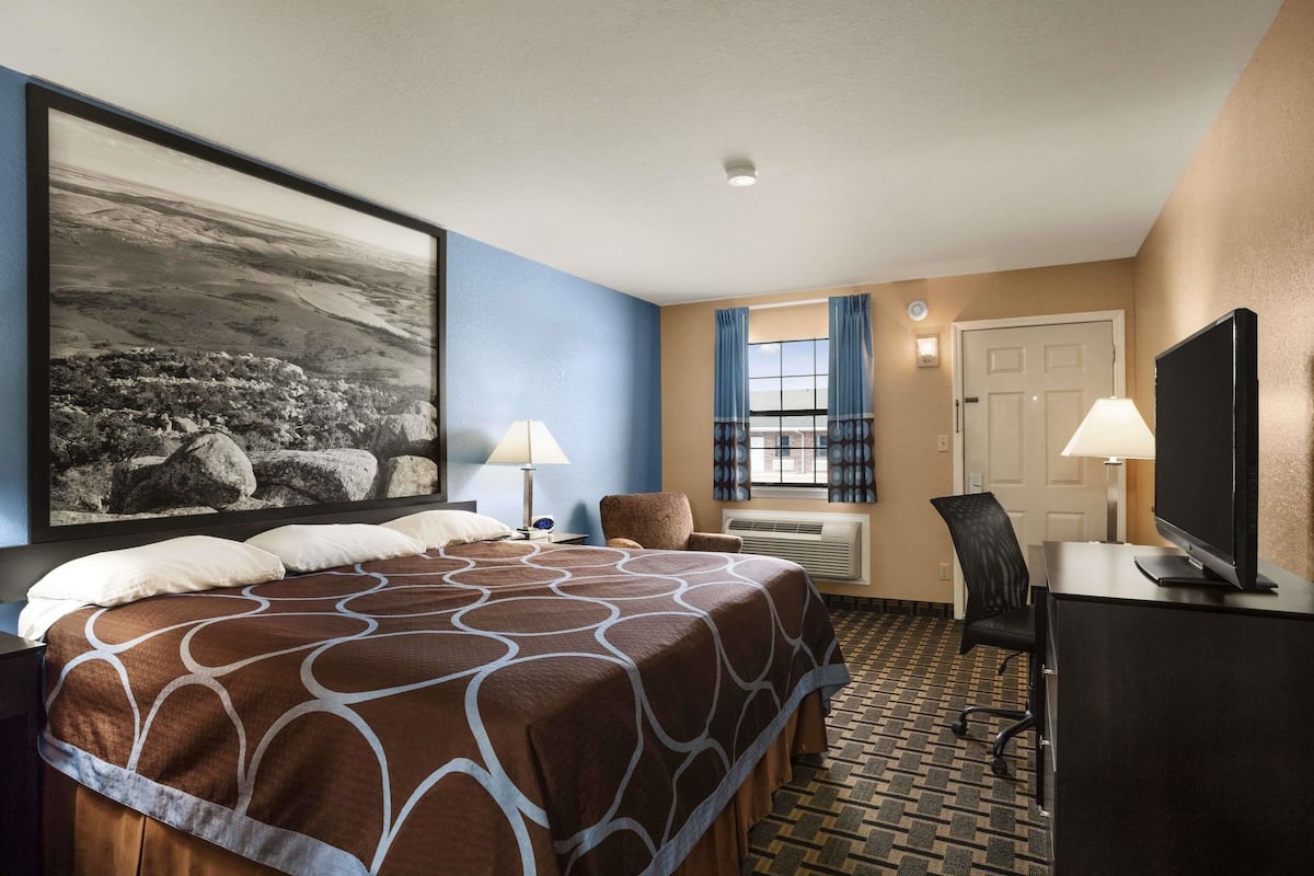 Quarto de hotel com uma cama grande, uma escrivaninha com televisão, uma cadeira, janela com cortinas e ar condicionado fixado na parede.