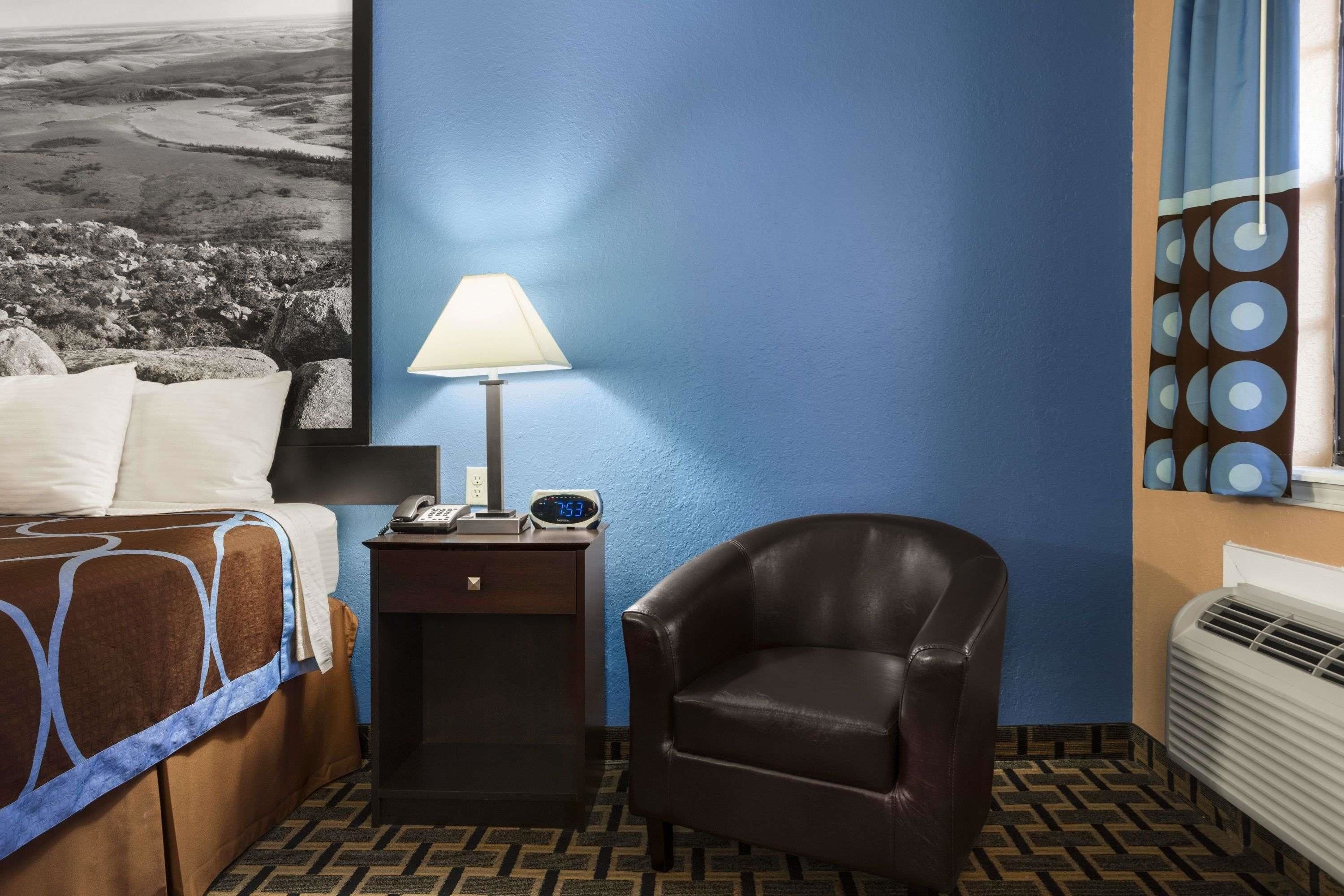 A hotel room with a bed, a nightstand with a lamp, a chair, a wall clock, and a window with curtains.