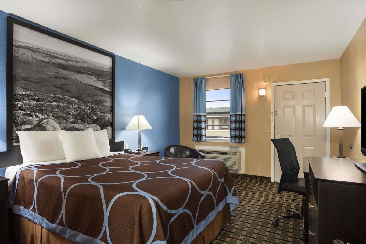 Quarto de hotel com uma cama grande, duas luminárias de cabeceira, uma escrivaninha com cadeira, uma janela com cortinas e ar condicionado fixado na parede.