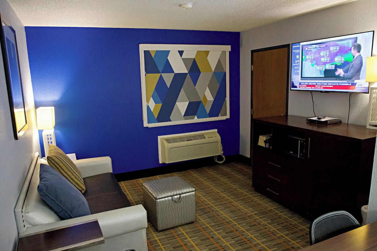 A hotel room with a blue accent wall, a large TV screen, a white sofa, a small ottoman, and a dark wooden cabinet.