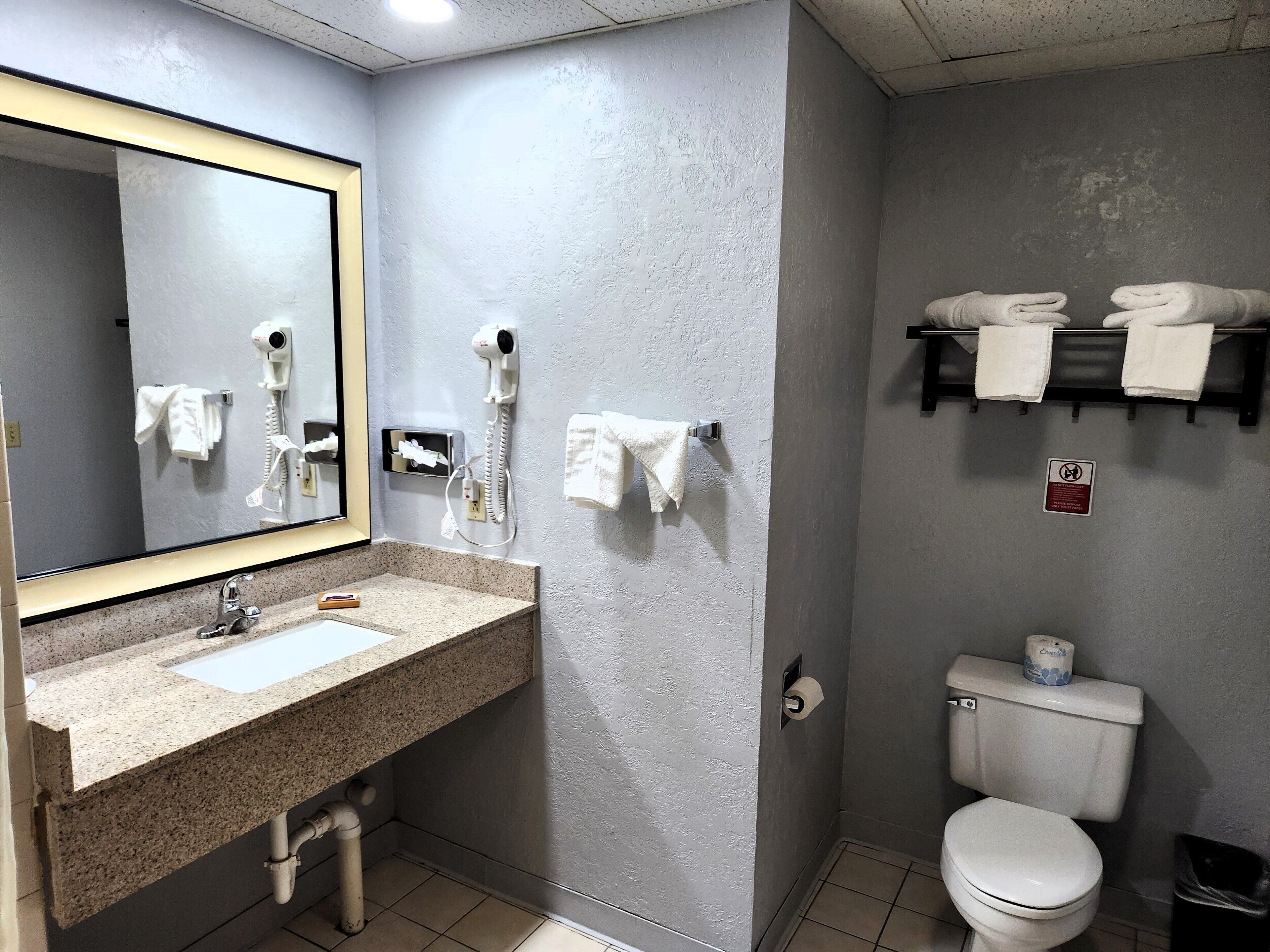 A bathroom with a granite countertop sink, a mirror, a hairdryer, a towel rack, a toilet, and a trash bin.