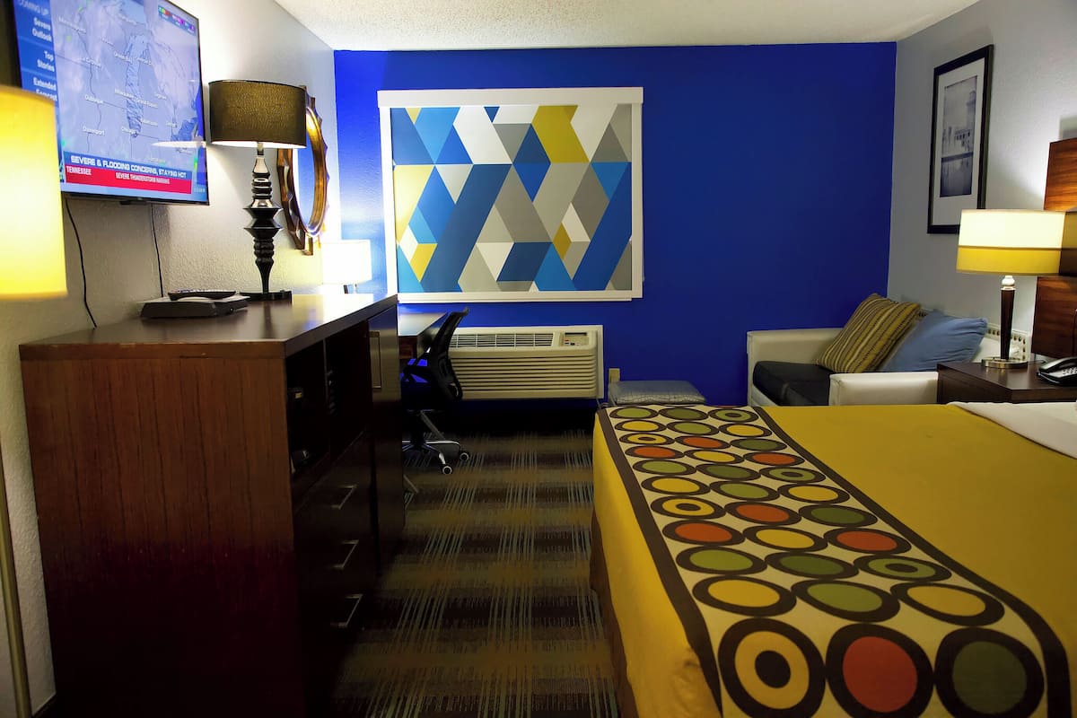 A hotel room with a bed, a desk, a chair, a TV, and a wall with a geometric painting.