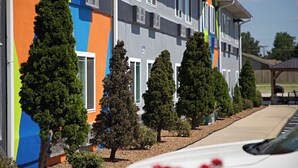 Un immeuble à plusieurs étages, doté d’une façade colorée, de nombreuses fenêtres et d’un espace de stationnement où se trouve une voiture blanche.