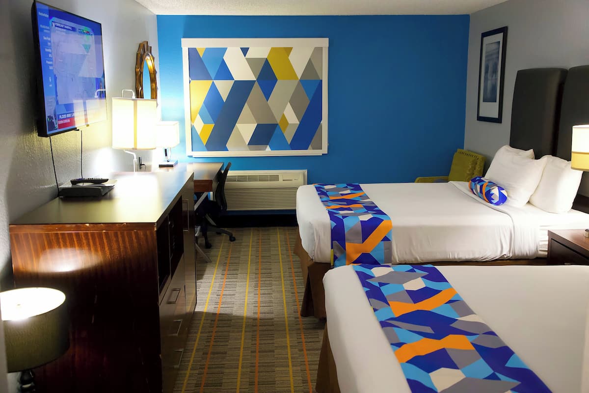 A hotel room with two beds, a desk, a TV, and a large geometric wall art piece.