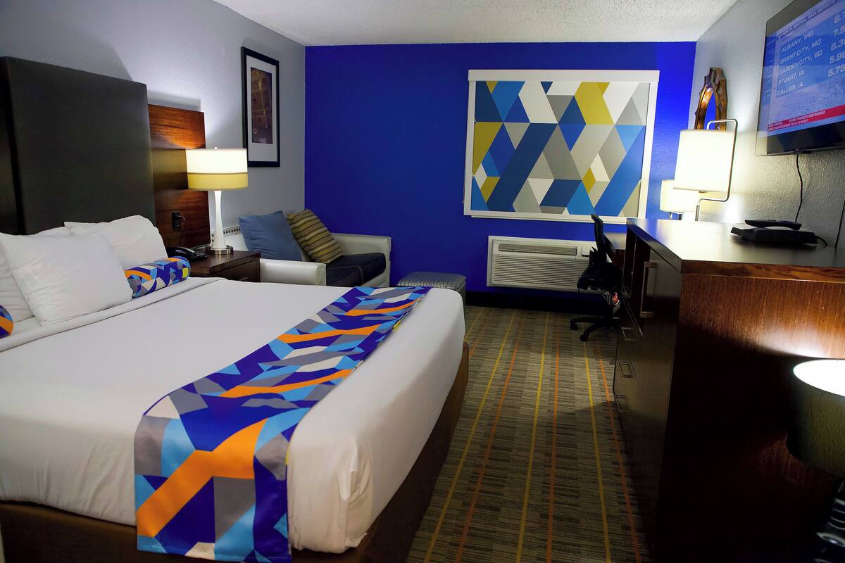 A hotel room with a large bed, a desk, a chair, a TV, and a wall with a geometric pattern.