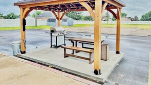 Un kiosque en bois équipé d’un barbecue et d’une table de pique-nique, installé sur une terrasse en béton.