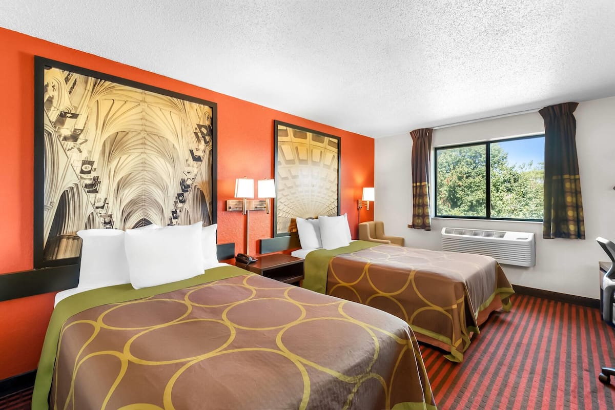 A hotel room with two beds, a large window with curtains, a desk with a chair, and a wall-mounted air conditioner.