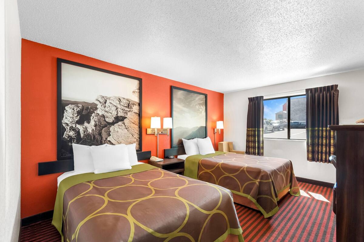 A hotel room with two beds, a large framed picture on the wall, a desk with a lamp, and a window with curtains.