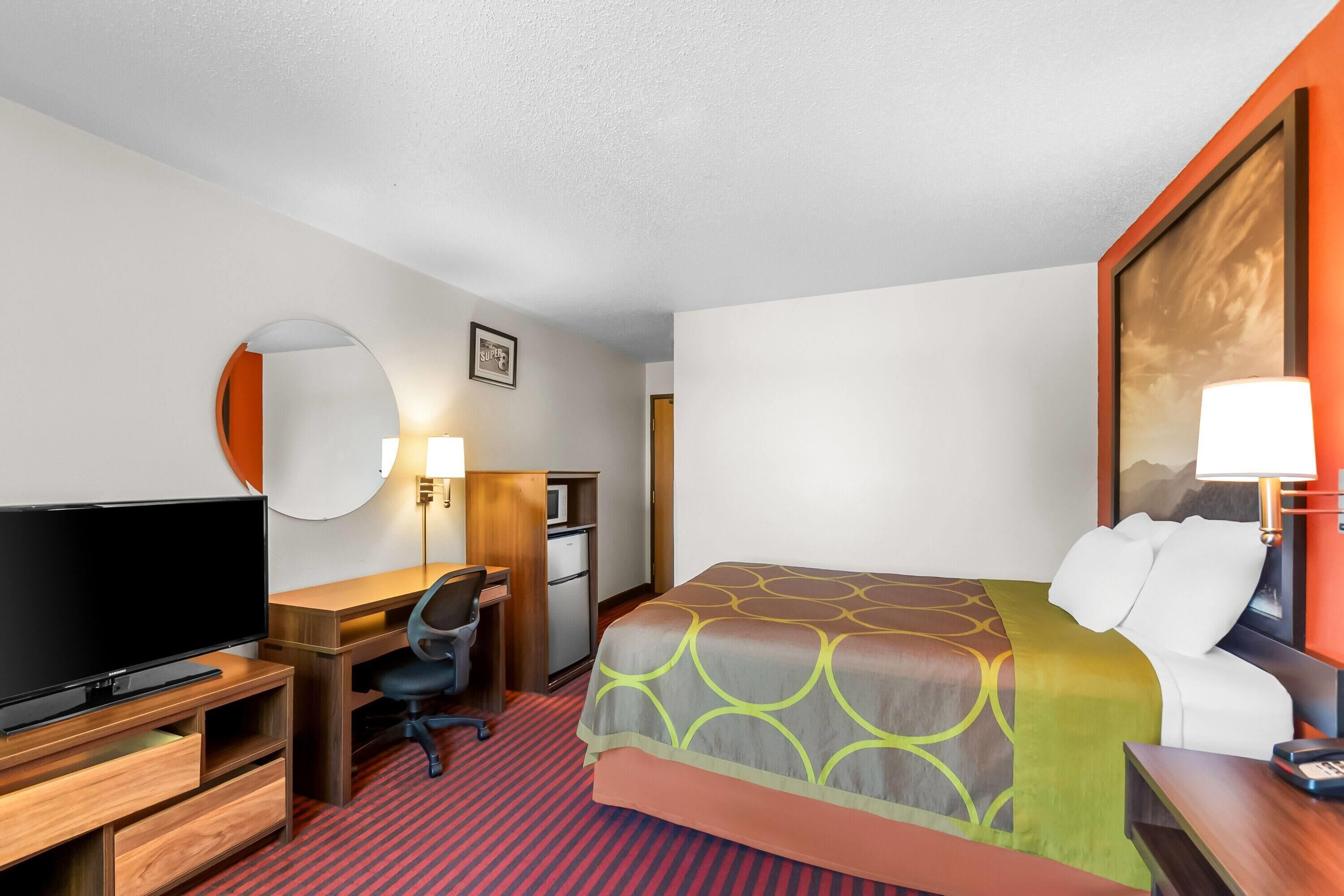 Habitación de hotel con cama, escritorio con silla, televisor, lámpara de escritorio y espejo.