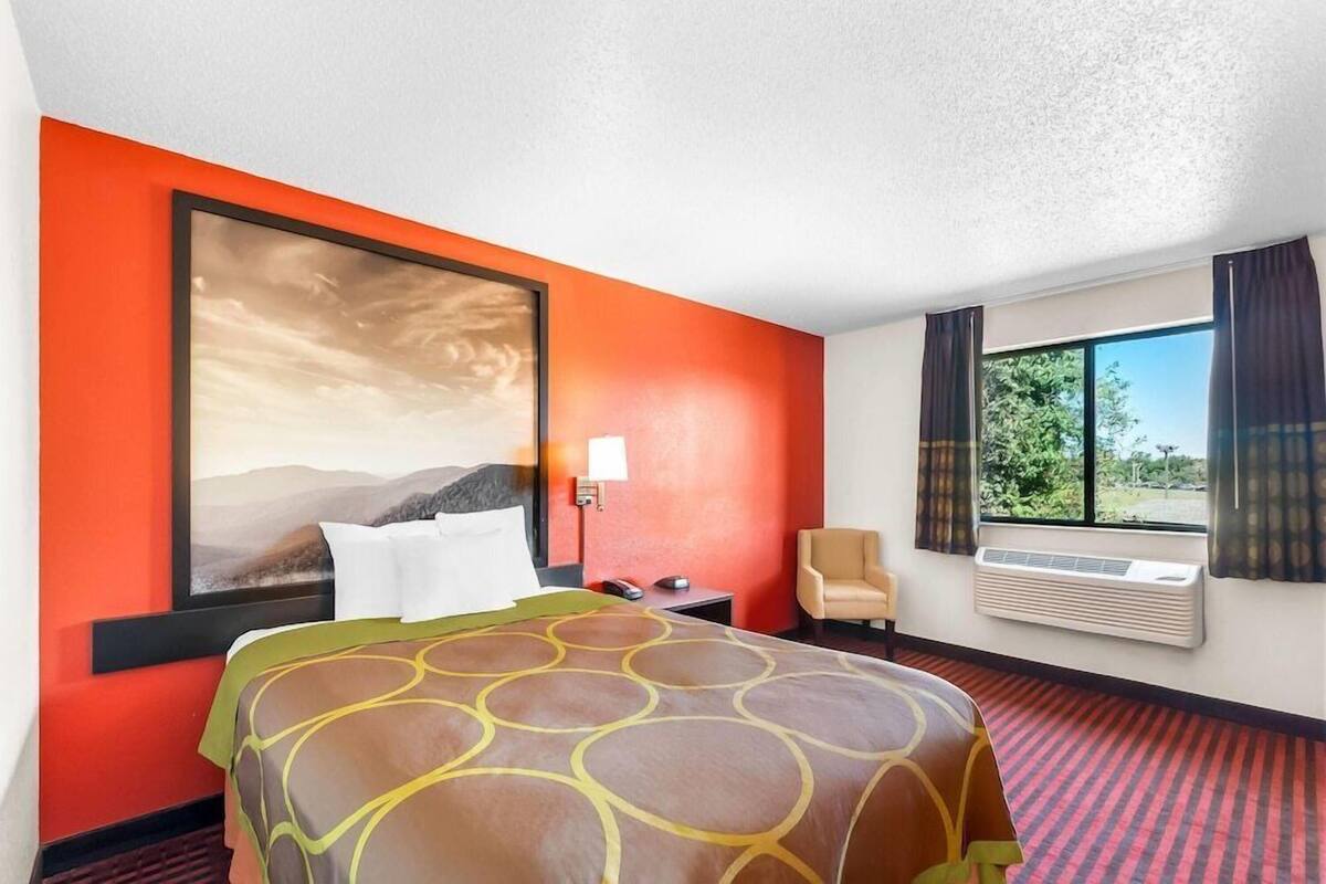 A hotel room with a large bed, a chair, a desk, a window with curtains, and a wall-mounted air conditioner.