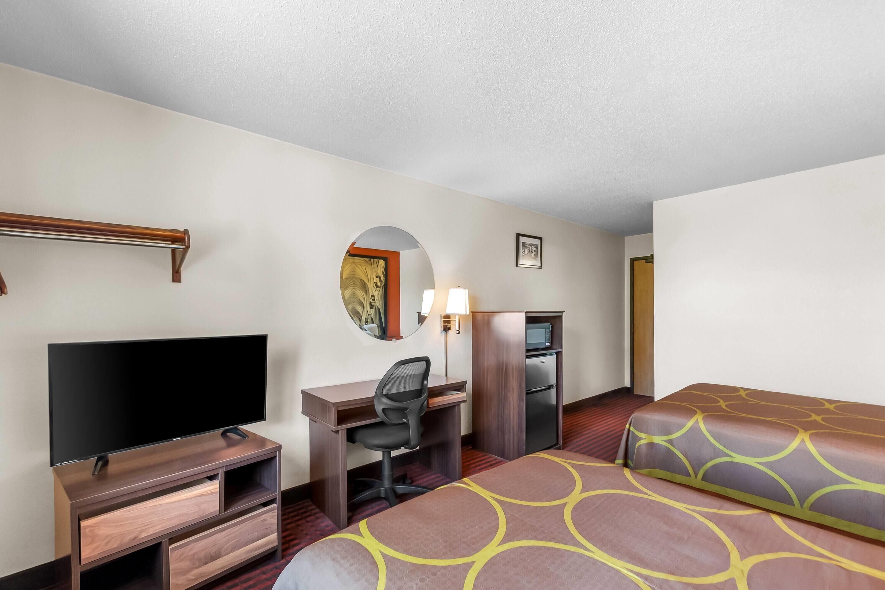A hotel room with a bed, a desk with a chair, a television, a refrigerator, a microwave, and a small mirror.