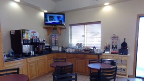 A small, well-lit kitchen with wooden cabinets, a coffee machine, and a television mounted above the counter. There are two round tables with chairs, and a window with blinds. - Super 8 by Wyndham Gillette (Gillette)
