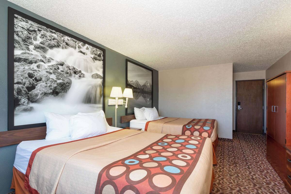 A hotel room with two beds, a large framed waterfall painting, a nightstand with a lamp, and a wardrobe.